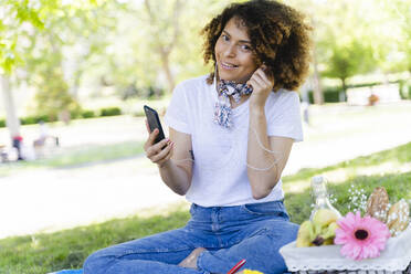 Entspannte Frau mit Mobiltelefon und Kopfhörern beim Picknick im Park - FMOF00696