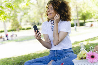 Glückliche Frau mit Mobiltelefon und Kopfhörern beim Picknick im Park - FMOF00697