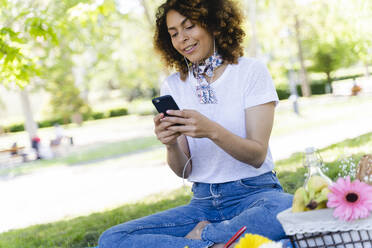 Entspannte Frau mit Mobiltelefon und Kopfhörern beim Picknick im Park - FMOF00698