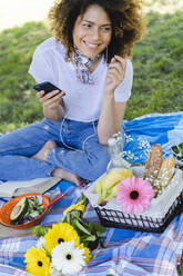 Entspannte Frau mit Mobiltelefon und Kopfhörern beim Picknick im Park - FMOF00700