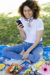 Entspannte Frau mit Mobiltelefon und Kopfhörern beim Picknick im Park - FMOF00701