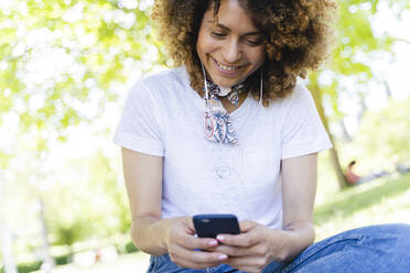 Smiling woman with cell phone and earphones in park - FMOF00702