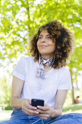 Lächelnde Frau mit Mobiltelefon und Kopfhörern im Park - FMOF00703