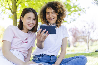 Zwei glückliche Frauen mit Handy und Kopfhörern im Park - FMOF00713