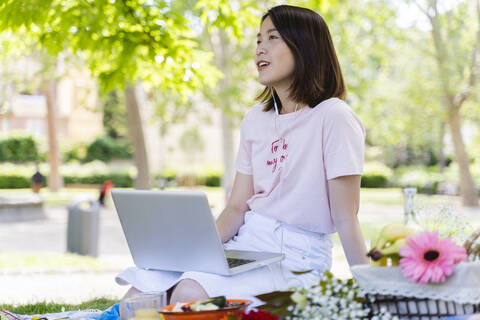 Junge Frau mit Laptop und Kopfhörern beim Picknick im Park, lizenzfreies Stockfoto