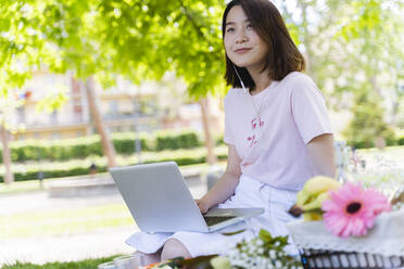 Junge Frau mit Laptop und Kopfhörern beim Picknick im Park - FMOF00725