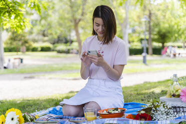 Entspannte Frau mit Mobiltelefon und Kopfhörern beim Picknick im Park - FMOF00726