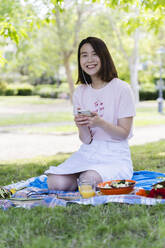 Glückliche junge Frau mit Handy und Kopfhörern beim Picknick im Park - FMOF00727