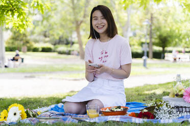 Glückliche junge Frau mit Handy und Kopfhörern beim Picknick im Park - FMOF00728