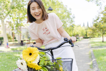 Porträt einer glücklichen jungen Frau mit Blumen und Fahrrad im Park - FMOF00741