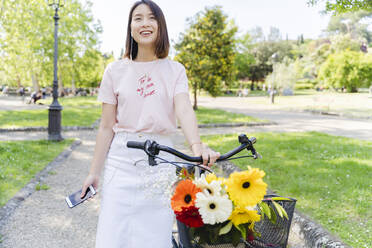 Lächelnde junge Frau mit Handy, Kopfhörer und Fahrrad im Park - FMOF00743