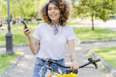Lächelnde Frau mit Handy, Kopfhörer und Fahrrad im Park - FMOF00744