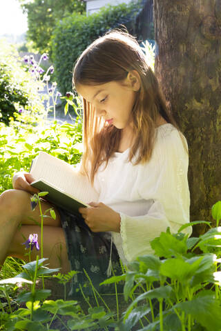 Kleines Mädchen lehnt an einem Baumstamm und liest ein Buch, lizenzfreies Stockfoto