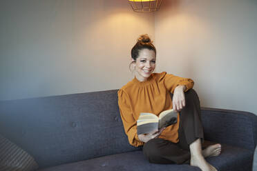 Portrait of smiling woman with book sitting on couch at home - PNEF01637