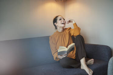 Lachende Frau mit Buch, die zu Hause auf der Couch sitzt - PNEF01638