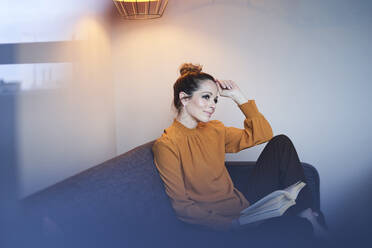 Smiling woman with book sitting on couch at home - PNEF01639