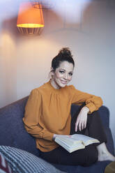 Portrait of smiling woman with book sitting on couch at home - PNEF01640