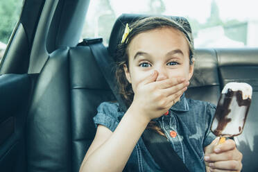 Mixed race girl eating ice cream bar in car back seat - BLEF07230