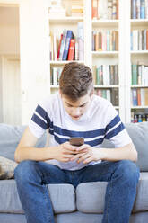 Teenage boy sitting on the couch at home using cell phone - LVF08111