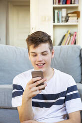 Portrait of smiling teenage boy sitting oin front of the couch using cell phone - LVF08113