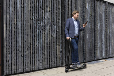 Businessman standing on pavement with with E-Scooter looking at cell phone - JOSF03281