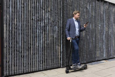 Geschäftsmann steht mit E-Scooter auf dem Bürgersteig und schaut auf sein Handy - JOSF03281