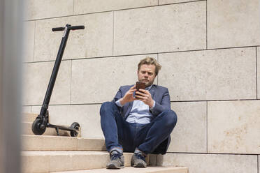 Businessman with E-Scooter sitting on steps using smartphone - JOSF03286