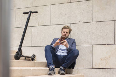 Geschäftsmann mit E-Scooter, der auf einer Treppe sitzt und ein Smartphone benutzt - JOSF03286
