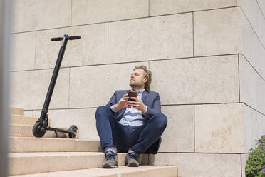 Geschäftsmann mit E-Scooter und Smartphone sitzt auf Stufen und entspannt sich - JOSF03288