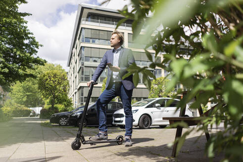 Businessman standing with E-Scooter and smartphone on pavement in the city - JOSF03290