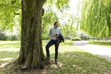 Geschäftsmann mit E-Scooter und Smartphone im Stadtpark - JOSF03297