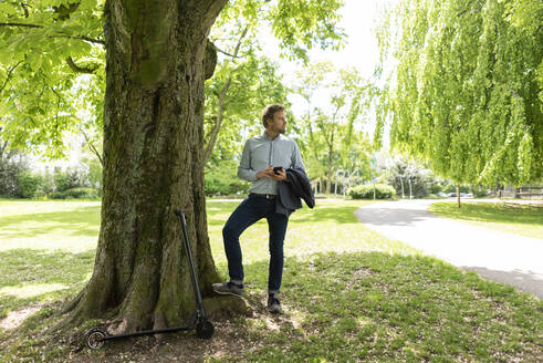 Businessman with E-Scooter and smartphone at city park - JOSF03297