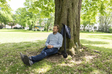Geschäftsmann sitzt auf einem E-Scooter in einem Park und lehnt sich entspannt an einen Baumstamm - JOSF03298