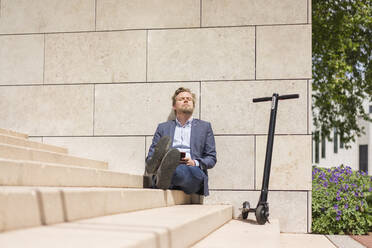 Businessman with E-Scooter and smartphone sitting on steps relaxing - JOSF03304