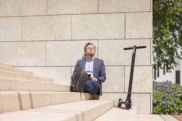 Geschäftsmann mit E-Scooter und Smartphone sitzt auf Stufen und entspannt sich - JOSF03304