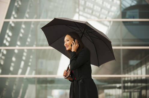 Asiatische Geschäftsfrau mit Regenschirm, die mit einem Handy telefoniert - BLEF07521