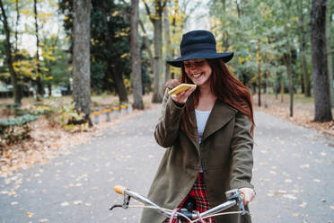 Young woman with long red hair on bicycle looking at smartphone in autumn park, Florence, Tuscany, Italy - CUF51428