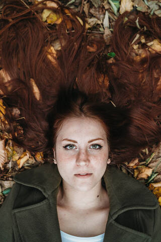 Junge Frau mit langen roten Haaren inmitten von Herbstblättern liegend, Porträt von oben, lizenzfreies Stockfoto