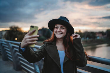 Junge Frau mit langen roten Haaren auf einer Fußgängerbrücke, die in der Abenddämmerung ein Smartphone-Selfie macht, Florenz, Toskana, Italien - CUF51431
