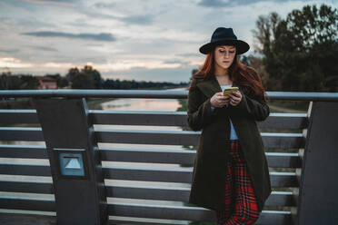 Young woman with long red hair leaning on footbridge looking at smartphone at dusk, Florence, Tuscany, Italy - CUF51432