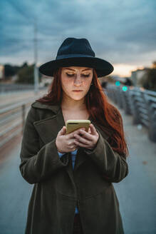 Junge Frau mit langen roten Haaren auf einer Fußgängerbrücke, die in der Abenddämmerung auf ihr Smartphone schaut - CUF51433