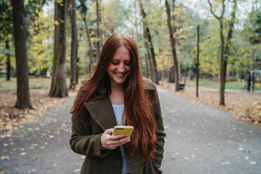 Young woman with long red hair looking at smartphone in tree lined autumn park, Florence, Tuscany, Italy - CUF51435