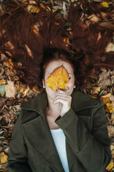 Young woman with long red hair lying amongst autumn leaves and covering face with autumn leaf, overhead portrait - CUF51437
