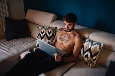 Mittlerer erwachsener Mann mit tätowierter Brust auf dem Sofa mit Blick auf den Laptop - ISF21436