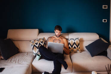 Mittlerer erwachsener Mann mit tätowierter Brust auf dem Sofa mit Laptop - ISF21437