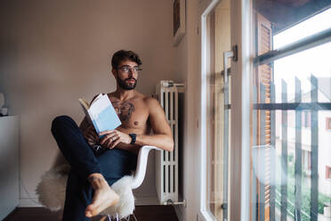 Mid adult man with tattooed chest sitting on chair looking out through window - ISF21441