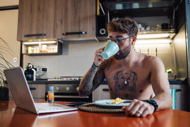 Mittlerer erwachsener Mann mit Tattoos, der Kaffee trinkt und auf einen Laptop schaut - ISF21444