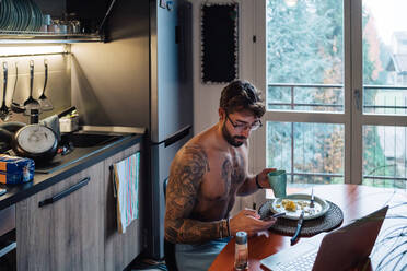 Mittlerer erwachsener Mann mit Tattoos, der Kaffee trinkt und auf sein Smartphone schaut - ISF21445