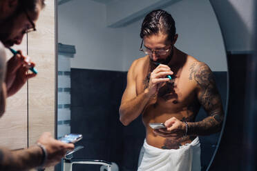 Mittlerer erwachsener Mann mit Tattoos, der sich die Zähne putzt und auf sein Smartphone im Badezimmerspiegel schaut, Spiegelbild - ISF21450