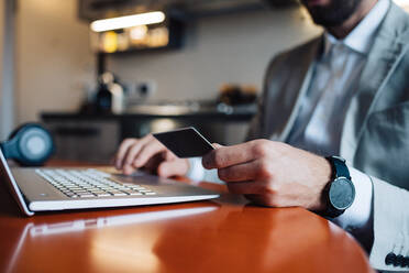 Mid adult man sitting at kitchen table with laptop and credit card, cropped - ISF21455